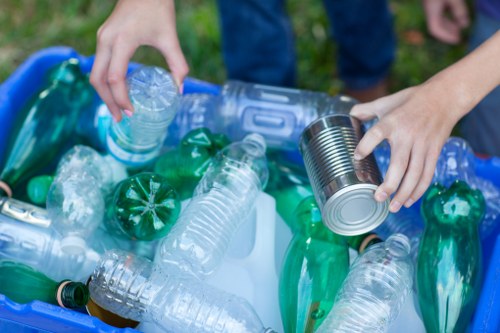 Close-up image of a person's hands, one holding a plastic water bottle and the other holding a tin can, both being placed into a blue recycling bin filled with various discarded plastic bottles, some with green and clear textures, and a few aluminum cans, set outdoors on a grassy area, suggesting a waste collection or rubbish removal activity in a local garden or yard, consistent with services offered by Garden Clearance Crystal Palace for rubbish disposal.