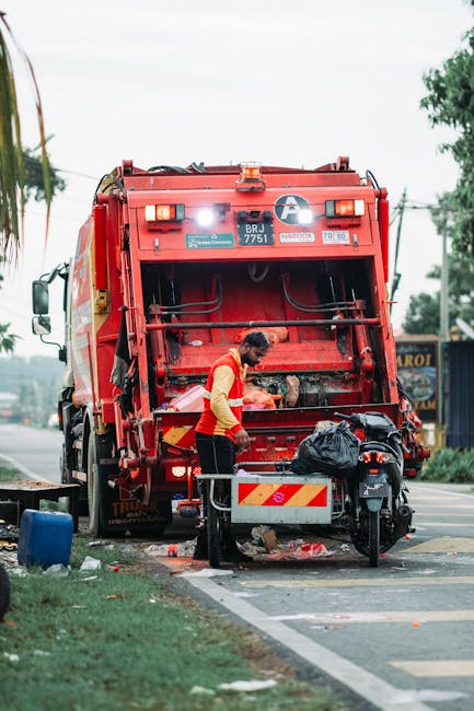 A street scene showing a waste collection truck operated by a waste management service, with the rear loading section open and partially rusted, revealing mechanical components used for compacting rub