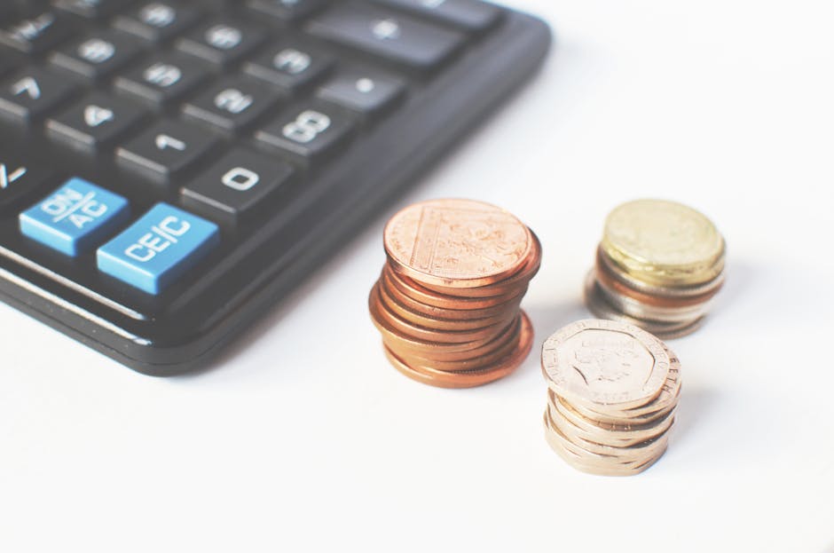 A black calculator with grey and blue buttons is partially visible in the top left corner of the image. In the foreground, there are three stacks of coins arranged on a white surface; the leftmost sta